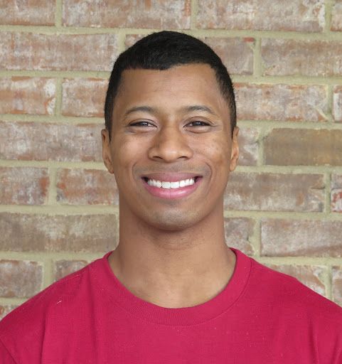 Patient Cory M of Just for Grins Orthodontics, young man wearing red shirt, smiling in front of brick wall